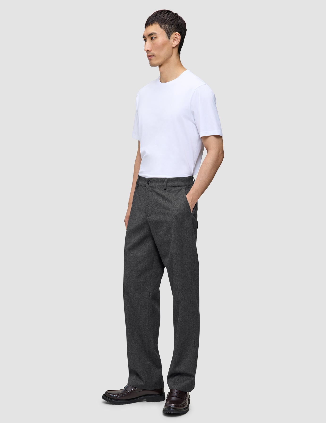En person i en hvit T-skjorte og mørkegrå Atelier Pants Relaxed-bukser står med den ene hånden i lommen.